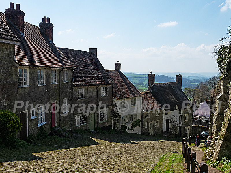The steep cobbled stree of Gold Hill with housing and the buttressed walls of the Precinct part of the ancient Shaftesbury Abbey, Dorset, England, UK, April 2023